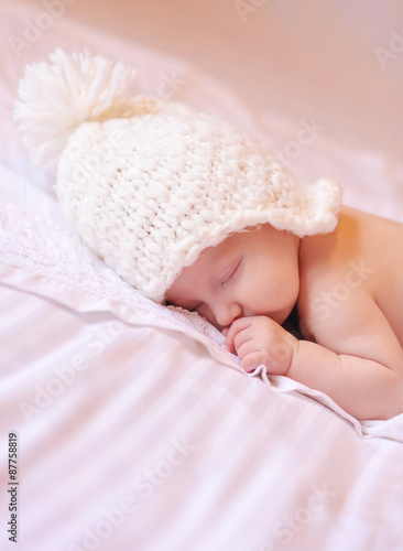 Little baby sleeping on the bed with white cover and knitted white hat on. Childhood. 