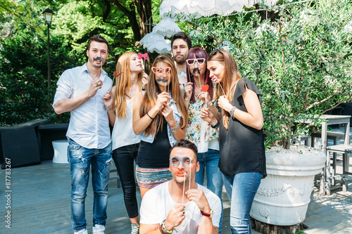 Party! A group of friends, three women and a man have fun at a party in a park with a mustache and fake glasses, joking and talking to each other  photo