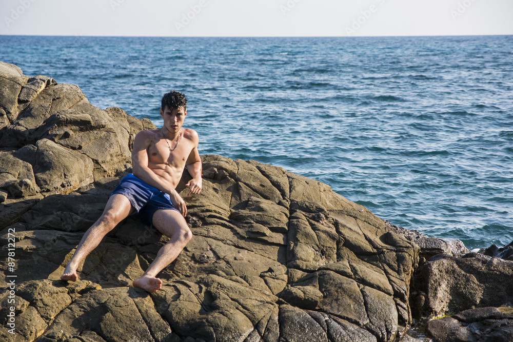 Young shirtless athletic man leaning on rock by water on ocean