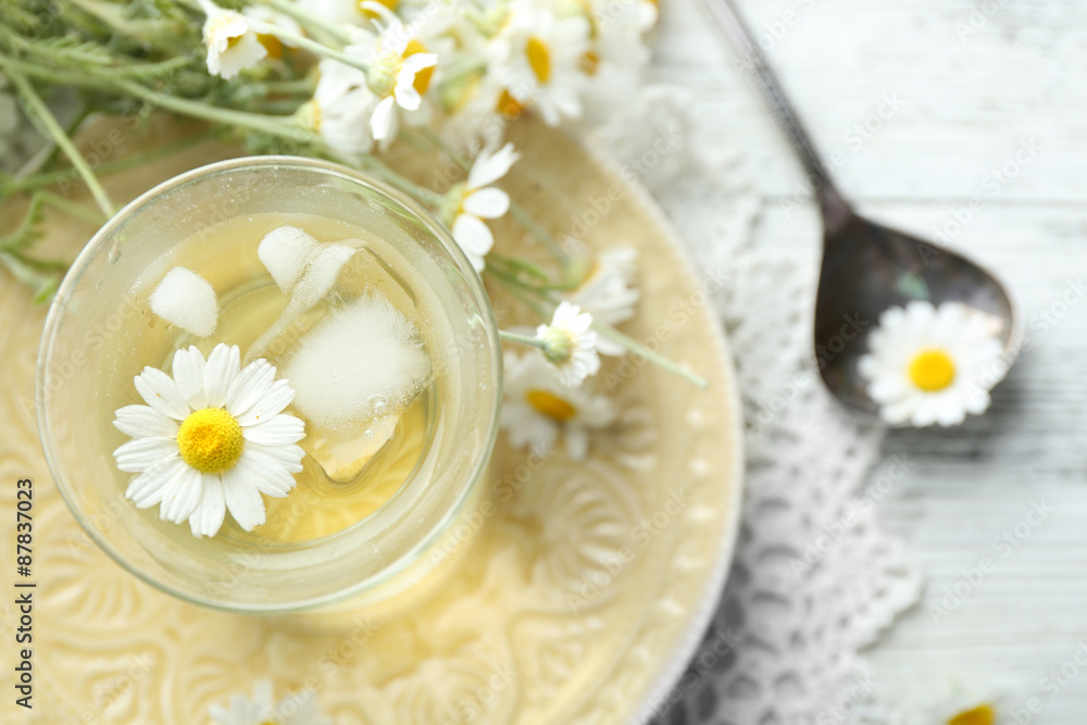 Glass of cold chamomile tea with ice cubes and chamomile flowers on color wooden background Glass of cold chamomile tea with ice cubes and chamomile flowers on color wooden background