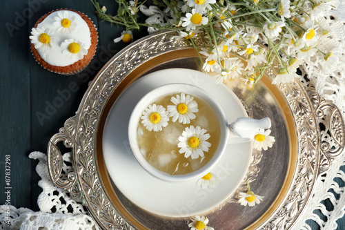 Cup of chamomile tea with chamomile flowers and tasty muffins on tray, on color wooden background