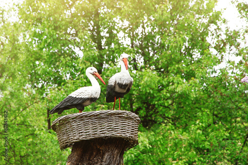 Decorative storks in wicker nest on trunk tree over green trees background