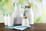 Pitcher, jars and glasses of milk on wooden table, on nature background