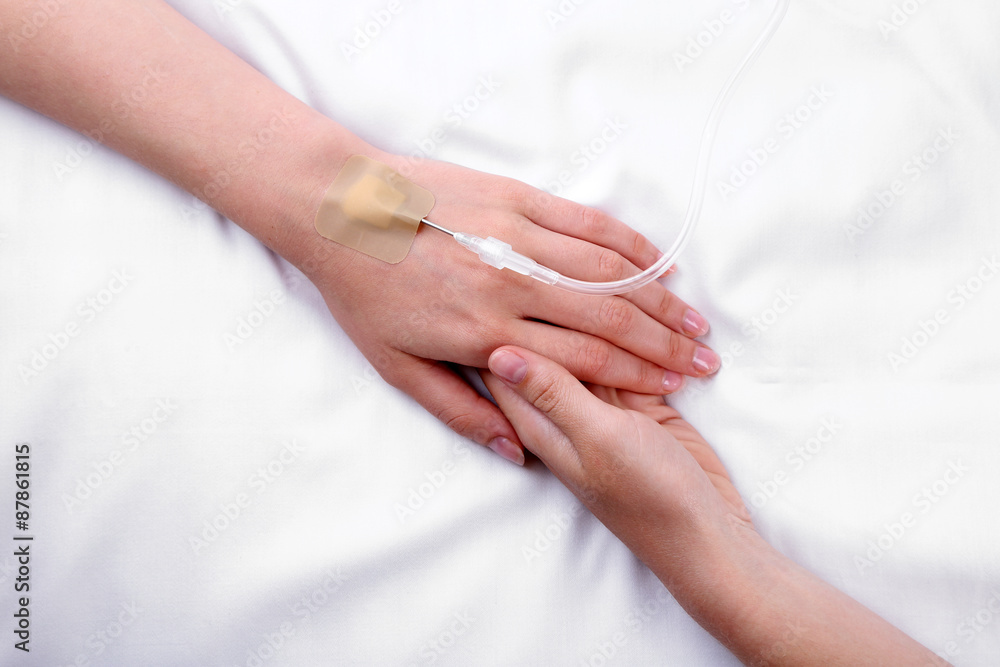 Female hands holding patient hand with dropper needle on bed close-up Female hands holding patient hand with dropper needle on bed close-up