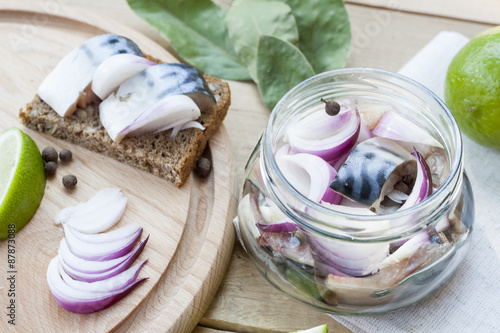 Slices of marinated mackerel with onion in a jar, lime, laurel and bread on wooden board, selective focus