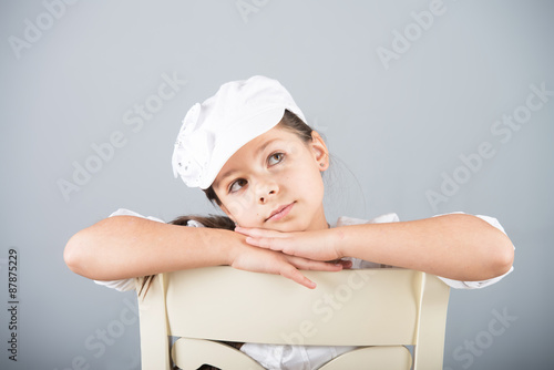 Portrait of a preteen girl in white cap sitting astride a chair and dreaming