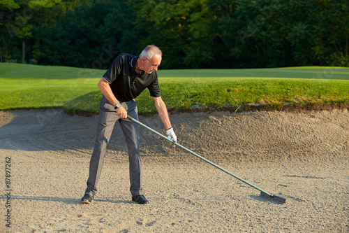 Obraz na plátně Senior golfer rake the the sand in a bunker