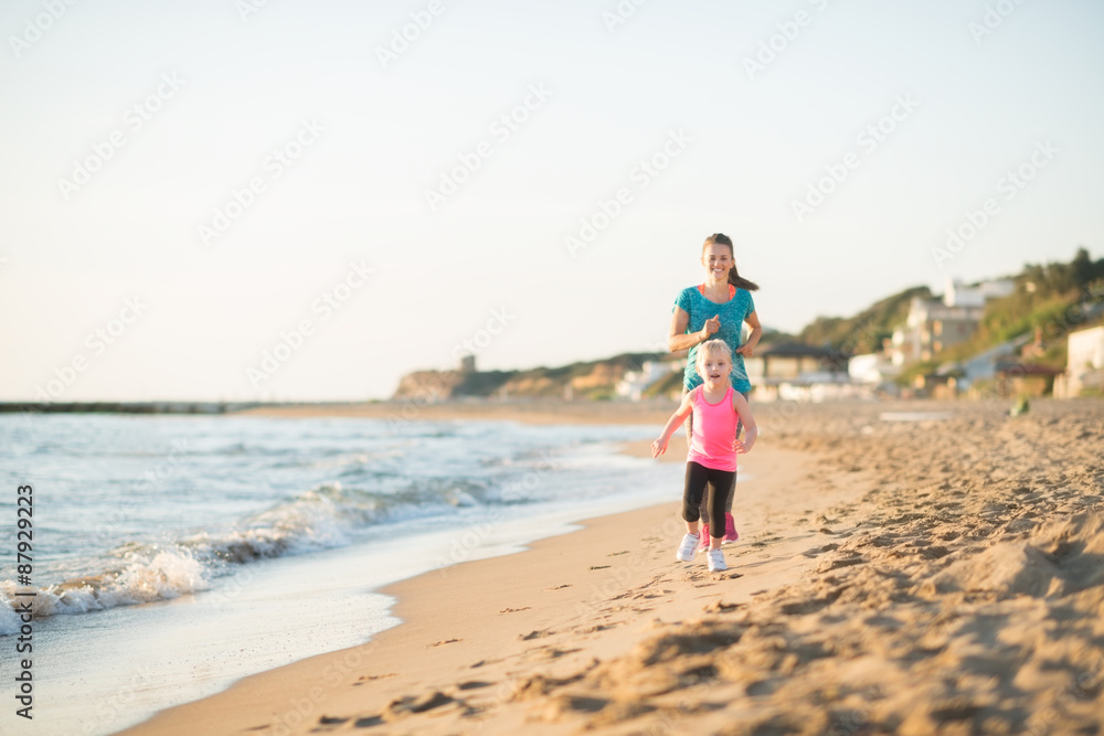 Mother and daughter running along water on beach at sunset