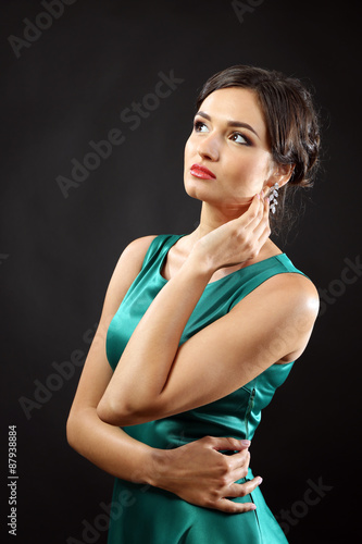 Elegant woman in emerald green dress with diamond earrings posing thoughtfully against a dark background, the female adult wears formal attire and makeup, exuding glamour and sophistication