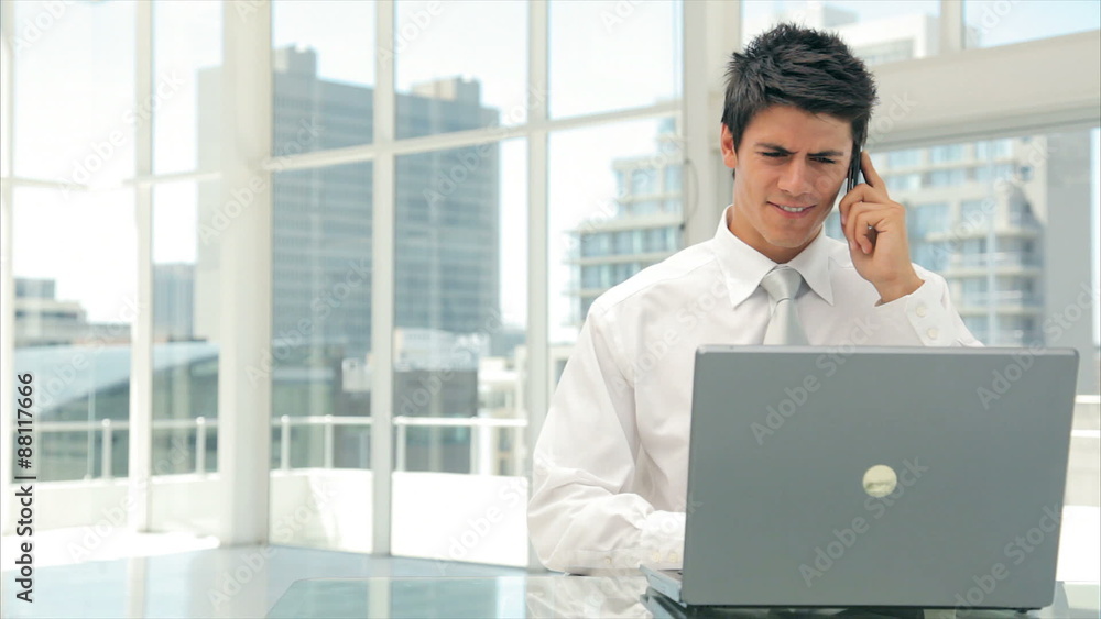 Lockdown shot of businessman answering smart phone while using laptop. Male executive is in formals. Smart well-dressed professional is sitting at desk in brightly lit office. Lockdown shot of businessman answering smart phone while using laptop. Male executive is in formals. Smart well-dressed professional is sitting at desk in brightly lit office.