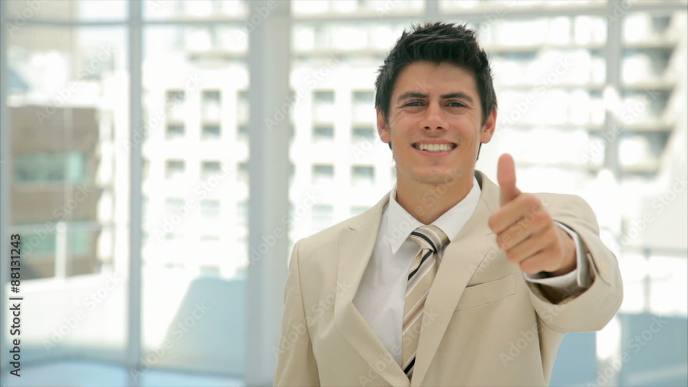 Panning shot of handsome businessman showing thumbs up. Happy young professional is in suit. Male executive is standing in brightly lit office. Panning shot of handsome businessman showing thumbs up. Happy young professional is in suit. Male executive is standing in brightly lit office.