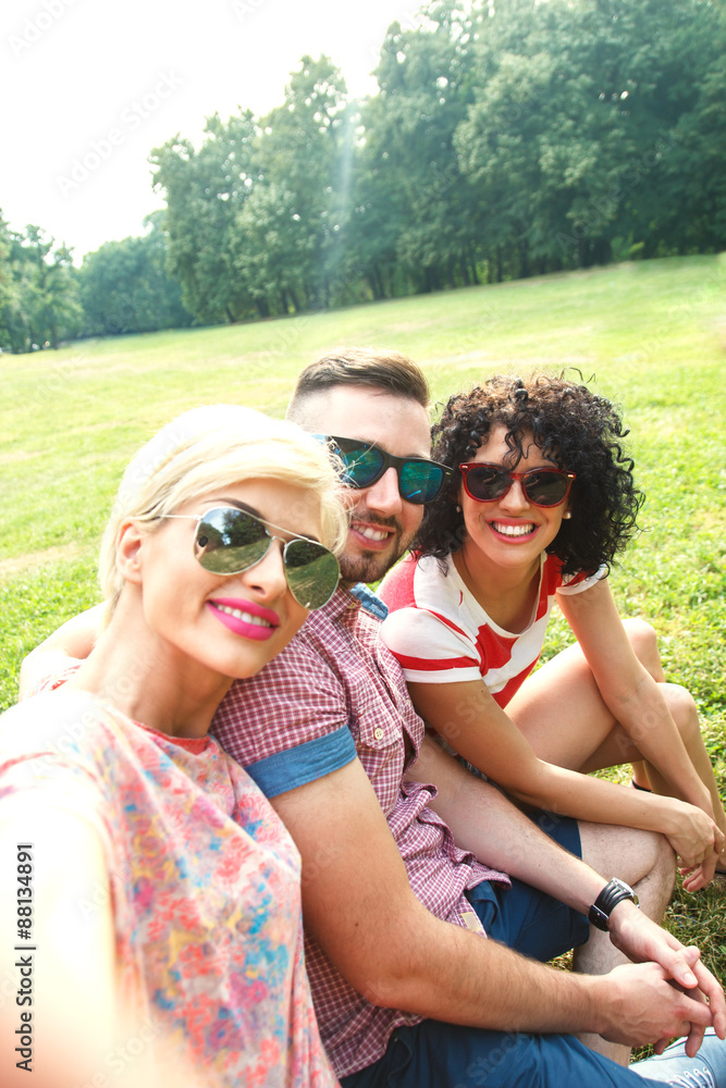 Three friends taking a selfie outdoors in park on sunny summer day. Two female and one male friends having fun photographing themselves on tablet.