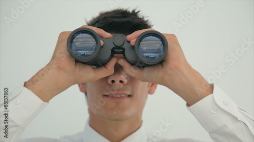 Tilt up image of young businessman using binoculars. Male executive is searching for opportunities. Professional is standing against wall.
