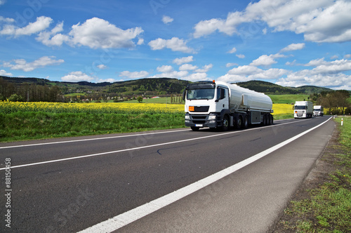 Obraz na płótnie White tanker and trucks driving along the asphalt road around the yellow flowering rapeseed field w salonie