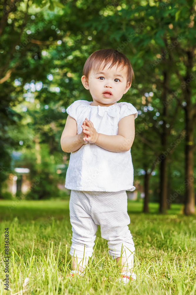 Portrait of cute adorable little Asian girl child baby one year old in white pants shirt, standing on field meadow grass on sunset playing Portrait of cute adorable little Asian girl child baby one year old in white pants shirt, standing on field meadow grass on sunset playing