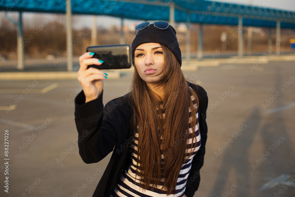 Outdoor fashion closeup portrait of nice pretty young hipster woman taking selfie sits on the tarmac at sunset.