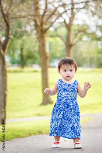 Portrait of a cute adorable little Asian girl child, one two years old, in blue dress and white shoes walking running in park outside on summer day, concept of first baby steps