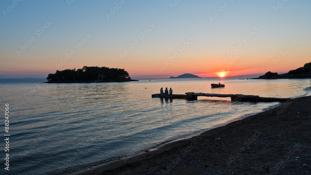 Girls sitting on a pier in front of small island at sunset
