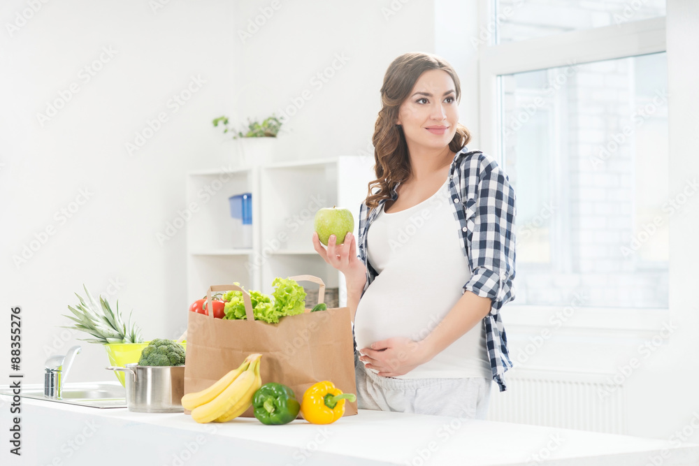 Beautiful pregnant woman in the kitchen with shopping bag and apple.