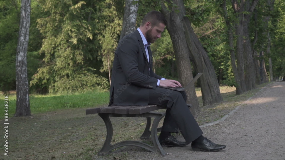 Businessman working on laptop, sitting on bench in park, pan shot.
Businessman working on laptop, sitting on bench in park, pan shot.