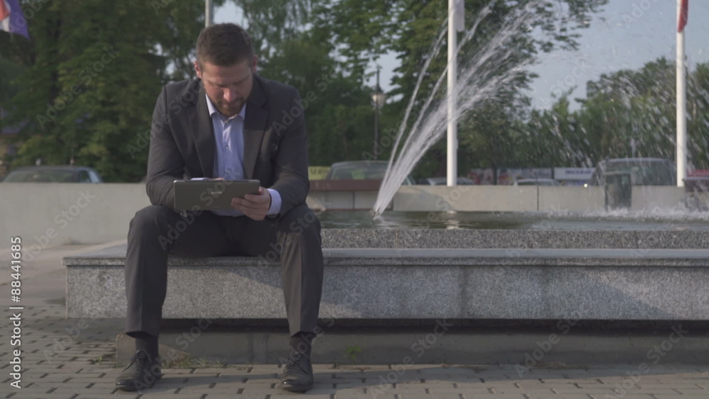Businessman sitting on wall, browsing tablet.
Businessman sitting on wall, browsing tablet.