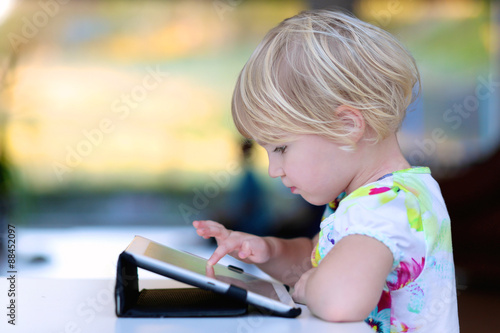 Happy little child, adorable blonde toddler girl enjoying modern generation technologies playing indoors using tablet pc with touchscreen