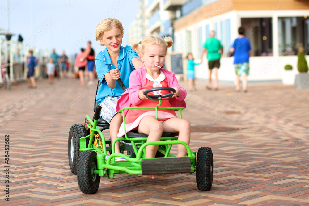 Happy kids enjoying active holidays on the beach. Sportive boy riding pedal car together with his cute toddler sister along the promenade on a summer day at sunset.