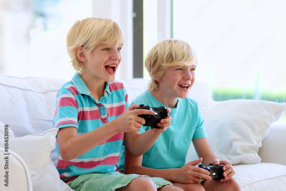 Two kids playing video game at home. Twin teenage brothers having fun after school day laughing and holding joysticks in hands sitting on white sofa in bright sunny living room with big windows Two kids playing video game at home. Twin teenage brothers having fun after school day laughing and holding joysticks in hands sitting on white sofa in bright sunny living room with big windows