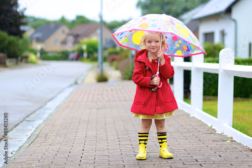 Happy little child, adorable blonde curly toddler girl wearing red duffle coat, bright yellow wellies and holding colorful umbrella walking on the street on a chilly autumn or spring day