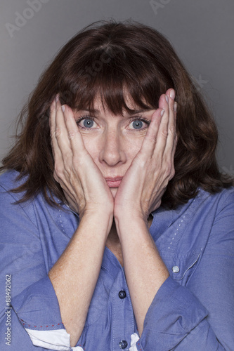 fear concept - attractive mature woman hiding her cheeks with both hands looking anxious and stressed out with eyes wide opened,closeup in studio shot