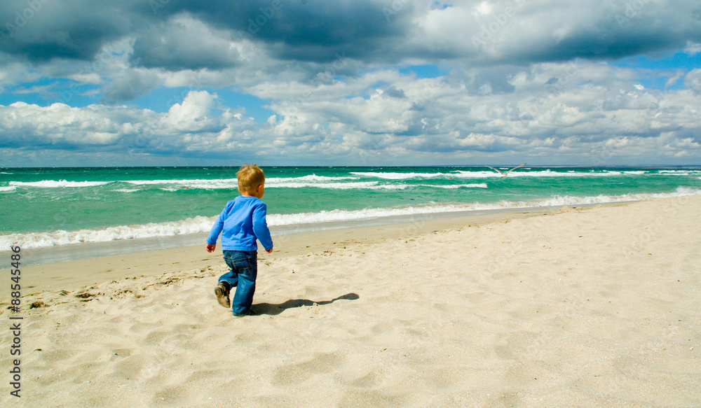 Little boy running along a beach on a cloudy sky background back view Little boy running along a beach on a cloudy sky background back view