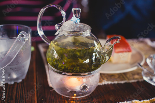 Beautiful warm picture of transparent teapot kettle with tasty green black tea with apple, lemon and ginger on a table with candles and with dessert in the background