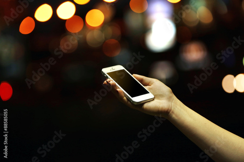 Female hand holding touch screen mobile phone on blurred night lights background