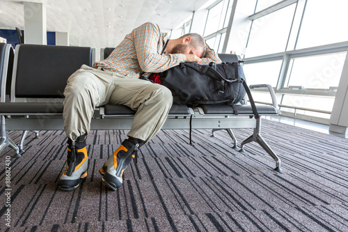 Man stuck at airport.
Image of bearded hippy style dressed person sleeping on his travel backpack inside airport waiting lounge sitting in black chair with heavy alpine boots on his legs