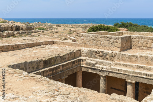 Looking down into a chamber of an ancient carved stone burial tomb at an archaeological site in Paphos, Cyprus with the blue Mediterranean Sea in the background.