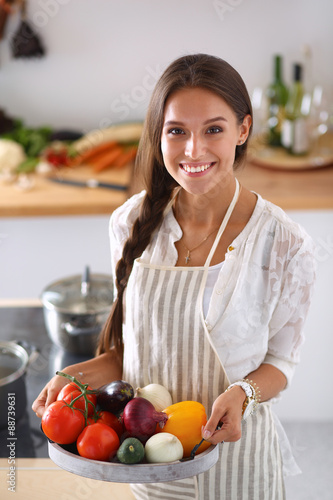 Smiling young woman holding vegetables standing in kitchen