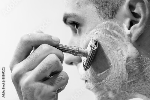 Handsome young man is shaving his face with razorblade and looking at the mirror photo