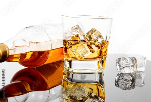 glass of whiskey near bottle and ice cubes on black table with reflection
