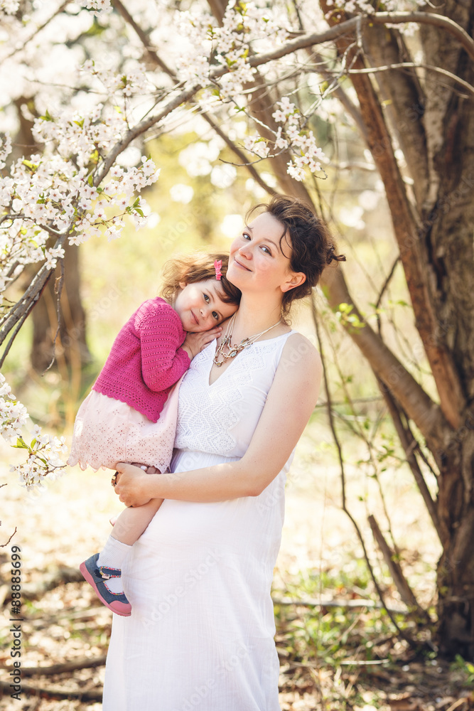 pregnant mother in white dress holding her daughter in pink clothes on spring summer day in park outside blooming cherry trees pregnant mother in white dress holding her daughter in pink clothes on spring summer day in park outside blooming cherry trees