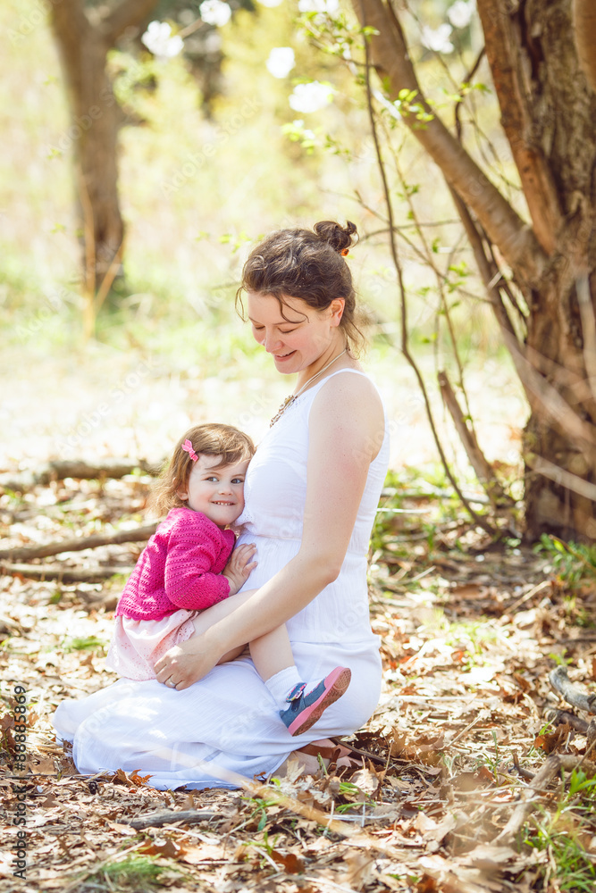 pregnant mother in white dress sitting on ground in park outside with daughter in pink clothes on her laps  on sunny spring summer day 