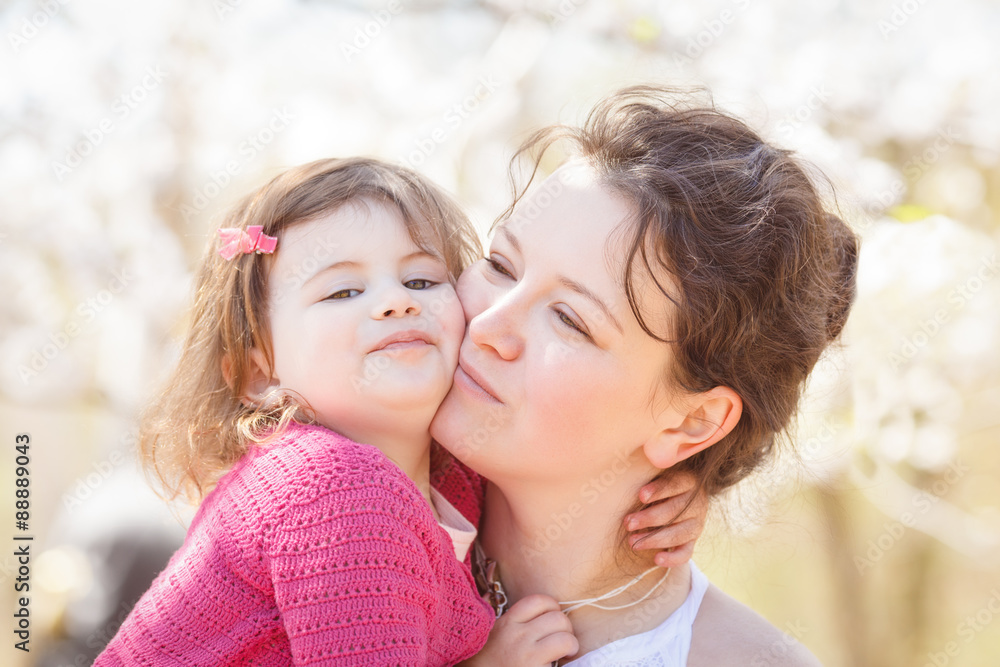 Close up portrait of caucasian mother in white dress holding hugging her daughter in pink clothes kissing her in cheek on sunny spring summer day in the park outside Close up portrait of caucasian mother in white dress holding hugging her daughter in pink clothes kissing her in cheek on sunny spring summer day in the park outside