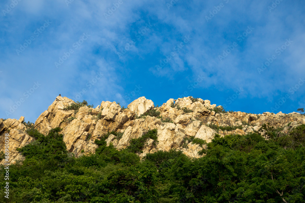 Stone or rock mountain with tree covering, blue sky and clounds