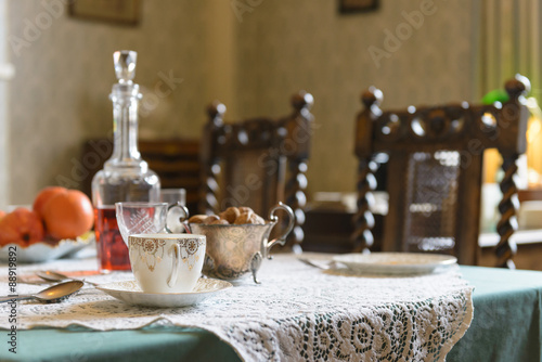 Vintage 1940s interior English table setting with teacup, nuts, wine, and oranges on a lace tablecloth.