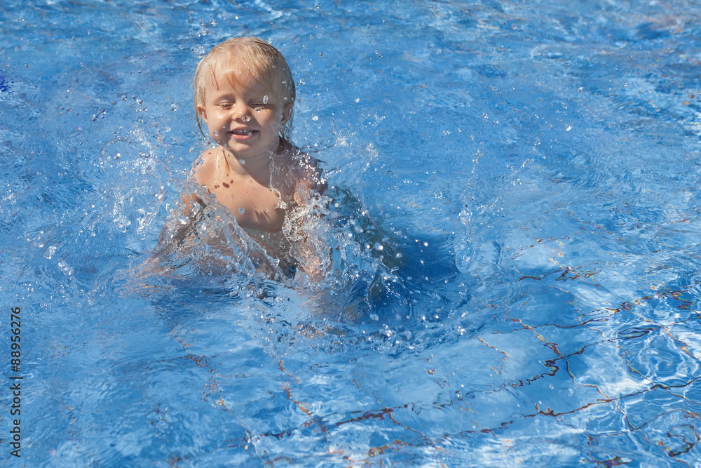 Happy smiling baby has fun jumping with splashes in blue water in pool before swimming lessons. Healthy lifestyle, active parents, and people water sports activity on summer family vacation with baby.