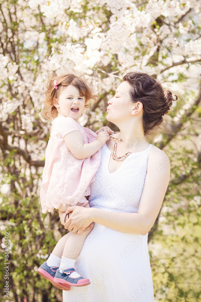 Portrait of caucasian pregnant mother in long white dress holding and kissing her daughter in pink clothes on spring summer day in park outside among blooming cherry trees 