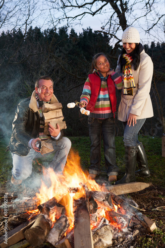 girl and her parents with a bonfire. The father is building up the fire with logs wilst the girl is holding marshmallows ready to roast with her mother. photo