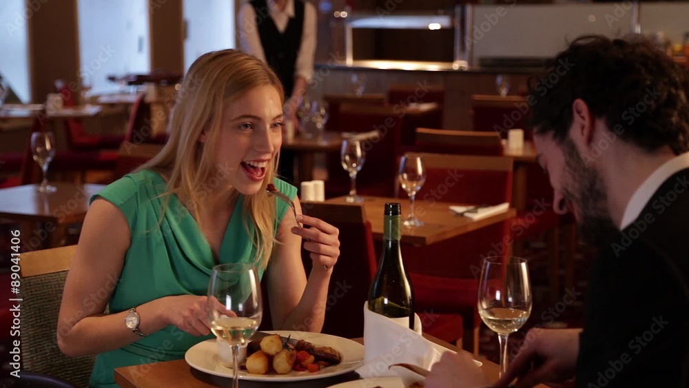 Young man and woman are sitting at a table in a restaurant. They are talking and eating and a waitress can be seen working in the background. Young man and woman are sitting at a table in a restaurant. They are talking and eating and a waitress can be seen working in the background.
