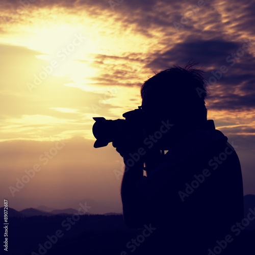 Professional photographer in jeans and shirt  takes photos with mirror camera on peak of rock. Dreamy landscape, orange  Sun at horizon © rdonar