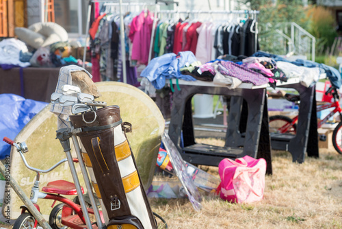 An old golf bag filled with clubs at a yard sale with a jumble of clotihng and toys in the soft background