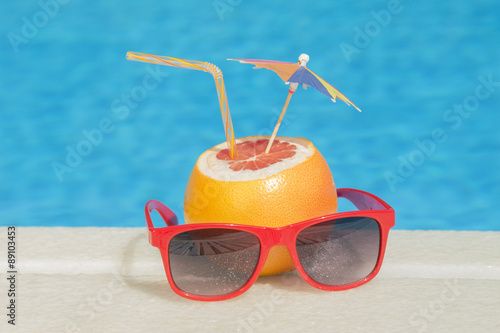 Grapefruit with drinking straw, umbrella and sunglasses at the pool edge 